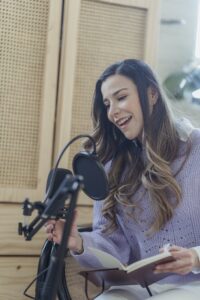 Content female in casual wear talking to professional microphone while recording audio record on chair with opened notebook in light room